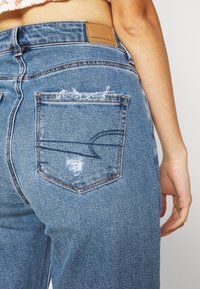 Jeans de mezclilla azul de talle alto con un ligero desgaste en los bolsillos traseros y una etiqueta de cuero marrón que muestra el nombre de la marca.