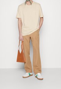 Beige bomull t-shirt, tan byxor, vita sneakers med gröna accenter, och en orange triangulär handväska. Enkelt och modernt design.