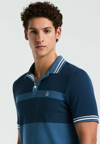 Men's polo shirt in dark blue, featuring textured fabric, striped collar in light blue and white, and a small logo on the chest.