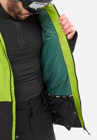 Veste de ski avec un extérieur noir et un intérieur vert fluo, dotée d'une poche zippée et d'une couche de base noire texturée en dessous.