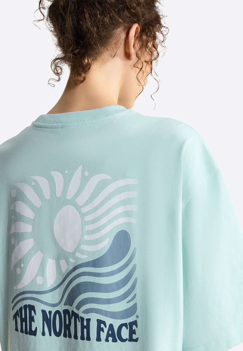 Person mit lockigem Haar, vom Betrachter abgewandt, trägt ein hellblaues Hemd mit einem stilisierten Sonnen- und Wellenmotiv sowie dem Schriftzug "THE NORTH FACE".