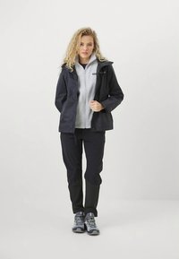 Mulher a usar uma impermeável preta sobre uma fleece cinza claro com fecho, calças pretas e sapatos de caminhada cinza, em pé contra um fundo branco liso.