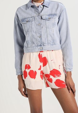 Chaqueta de denim azul claro con un diseño corto, dos bolsillos en el pecho y botones plateados, combinada con pantalones cortos de crema con patrones de manchas rojas.