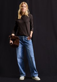 Schwarzes geripptes Langarmoberteil, hellblaue weite Jeans, hält eine braune Wildledertasche. Weiße Sneakers mit strukturierten Sohlen.