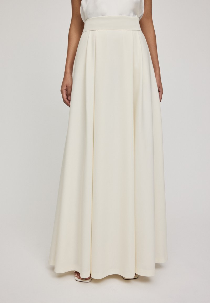 Jupe longue et fluide blanche avec une taille haute, fabriquée en tissu lisse. Elle présente des plis et un ourlet large, créant une silhouette élégante.