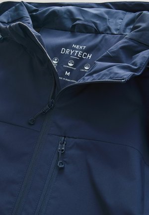 Tamsiai mėlyna vandeniui atspari striukė su lygiu audinio paviršiumi, turinti priekinę užtrauktuką ir dvi užtrauktuku užsegamas kišenes. Etiketėje matomas užrašas „NEXT DRYTECH“.