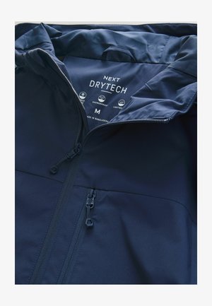 Giacca impermeabile blu navy con una texture liscia del tessuto, dotata di una cerniera frontale e due tasche con cerniera. L'etichetta mostra il marchio "NEXT DRYTECH".