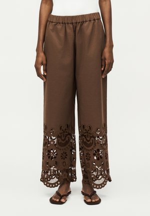 Brune bukser med vidde ben og elastik i taljen samt indviklede blomstrede blondeudskæringer ved kanten, båret med mørkebrune sandaler med åben tå.