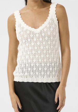 Femme portant un haut blanc en crochet sans manches avec des bords festonnés et une jupe noire sur un fond uni.