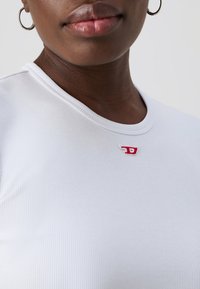 White ribbed T-shirt with a round neckline featuring a small red embroidered logo at the chest. Smooth texture, fitted design.