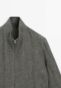 Grå zip-up jakke med høj krave, struktureret overflade og et subtilt ternet mønster, med et slankt og minimalistisk design.