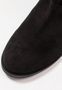 Zbliżenie na czarny zamszowy czubek buta o zaokrąglonym kształcie i gładkiej czarnej podeszwie na białym tle.