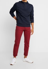 Man in een marineblauwe zip-up sweatshirt, opgerolde rode broek en witte sneakers, staand met één hand op de heup tegen een effen achtergrond.