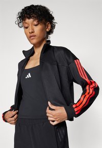 Zwarte sportjack met ritssluiting en rode strepen op de mouwen. Matte textuur met een aansluitend ontwerp, gedragen over een zwart tanktop met een logo.