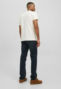 Beige bomullströja med korta ärmar, kombinerad med mörka denimjeans. Bruna mockaskor fulländar looken. Enfärgad bakgrund.