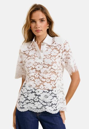 Woman wearing a white floral lace short-sleeve blouse with a pointed collar and decorative buttons, paired with dark blue jeans.