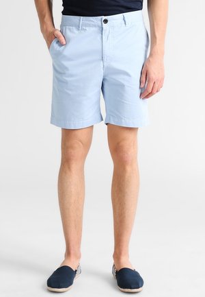Mand iført lyseblå shorts, marineblå slip-on sko og en sort trøje med den ene hånd i lommen, står på en ensfarvet hvid baggrund.