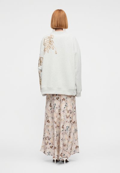 Γυναίκα με κοντά κόκκινα μαλλιά που φοράει υπερμεγέθη λευκό φούτερ με κεντήματα λουλουδιών και μακριά φλοράλ φούστα, η οποία στέκεται γυρισμένη προς τα πίσω.