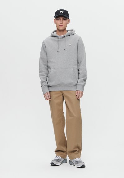 Gray hoodie with a front pocket, drawstring hood, worn over beige pants. Black cap with logo, gray athletic shoes with a white and black design.