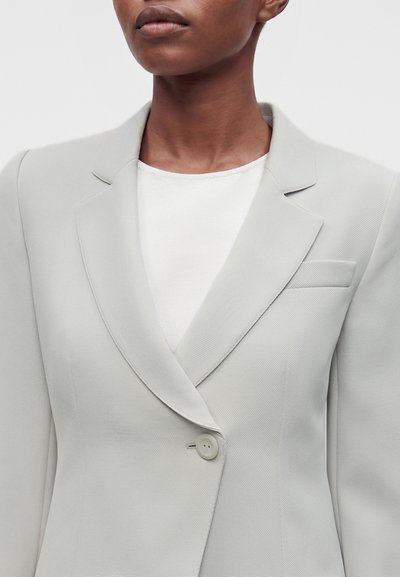 Personne portant un blazer gris clair sur mesure à un seul bouton par-dessus un haut blanc, montrée des épaules à la poitrine sur fond uni.