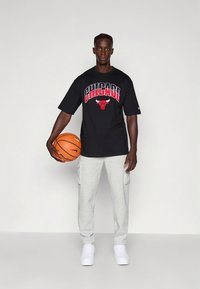 T-shirt preta de algodão com a palavra "CHICAGO" e o logotipo de um touro em vermelho, combinada com calças de fato de treino cinza e ténis brancos, segurando uma bola de basquetebol.