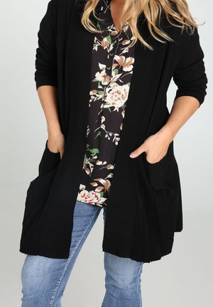 Frau trägt eine schwarze offene Strickjacke über einer geblümten Bluse mit blauen Jeans, Hände in den vorderen Taschen der Strickjacke.
