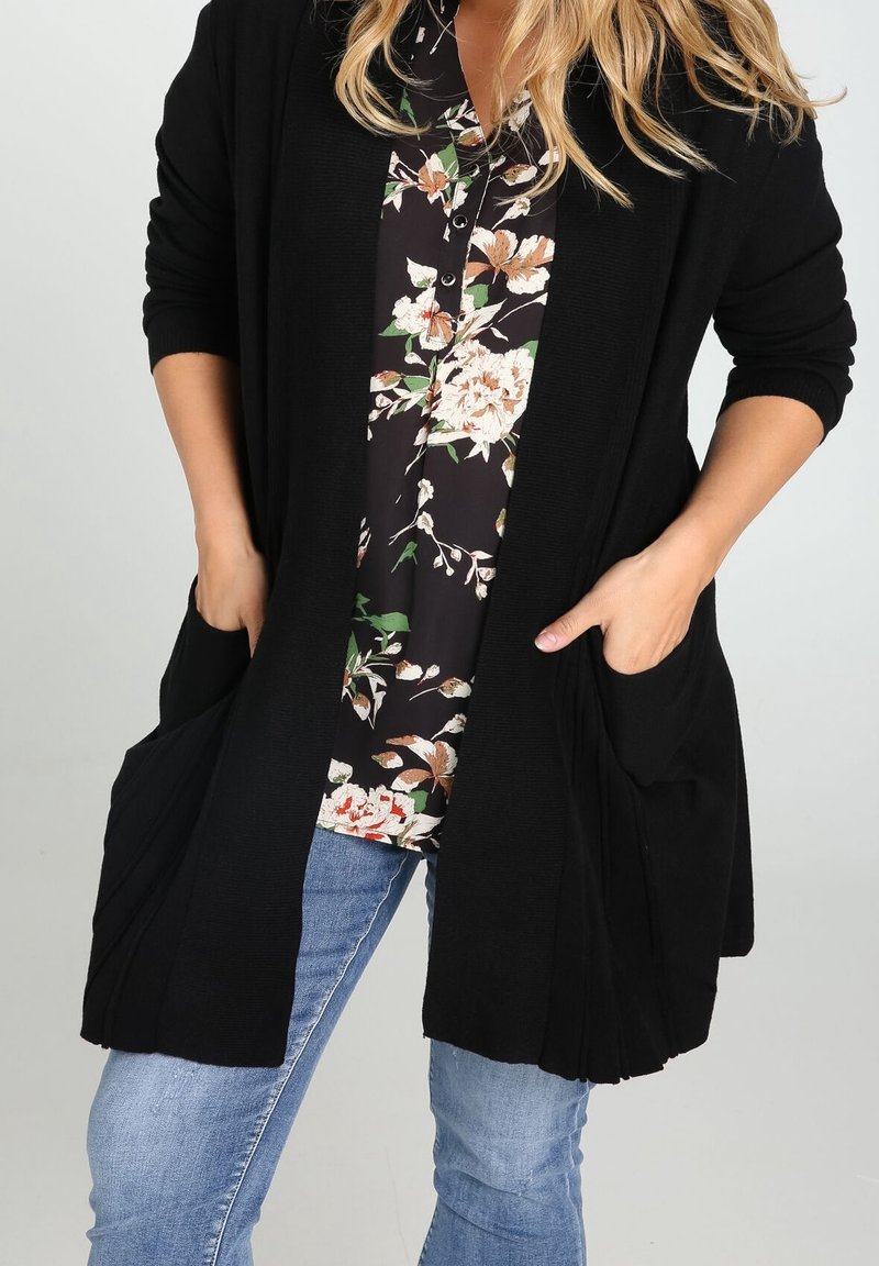 Femme portant un cardigan noir ouvert par-dessus un chemisier fleuri avec un jean bleu, les mains glissées dans les poches avant du cardigan.