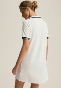 Biała sukienka polo z krótkim rękawem, z morskimi paskami na kołnierzyku i rękawach. Wykonana z fakturowanej tkaniny, o długości do kolan.