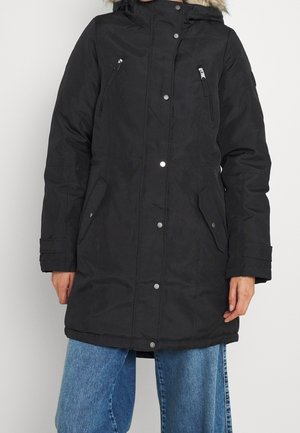 Zwarte parka met capuchon, met drukknoopsluiting aan de voorkant, ritssluitingen op de borstzakken, klepzakken op de heupen, gedragen met een blauwe spijkerbroek door een persoon tegen een witte achtergrond.