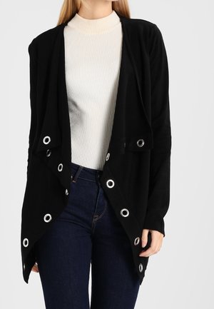 Femme portant un cardigan noir ouvert devant avec des œillets argentés sur un col roulé côtelé blanc et un jean bleu foncé.
