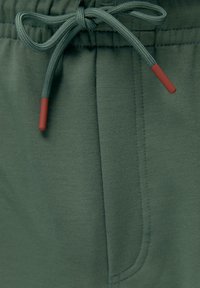 Zelené fleece nohavice so sťahovacím pásom, hladkou textúrou a červenými akcentmi na šnúrkach.