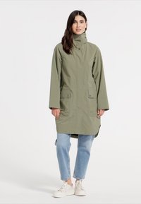 Groene waterdichte jas met een hoge kraag, twee voorzakken en een ontspannen pasvorm. Gecombineerd met lichtblauwe jeans en witte sneakers.