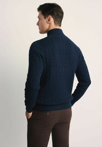 Granatowy sweter w splot kabla z prążkowanym kołnierzem i mankietami, o dopasowanym kroju. Nosi się go na brązowych spodniach, z widocznym teksturowanym detalem z tyłu.