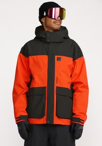 Veste de neige orange et noire avec capuche, grandes poches et fermeture éclair. Fabriquée en matériau imperméable et dotée de poignets ajustables.