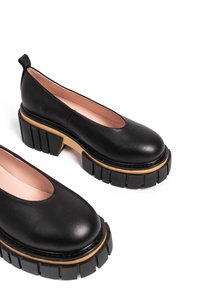 Scarpe slip-on in pelle nera con punta arrotondata, suola chunky nera con dettagli in legno e una finitura liscia e testurizzata.