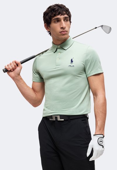 Homme en polo vert et pantalon noir tenant un club de golf sur l’épaule, avec un gant de golf blanc, regardant sur le côté.