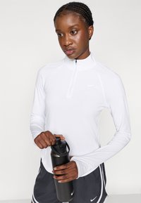 Λευκή αθλητική μπλούζα με μακριά μανίκια και φερμουάρ μέχρι το ένα τέταρτο, συνδυασμένη με μαύρη βερμούδα. Το άτομο κρατάει ένα μαύρο μπουκάλι νερού. Μινιμαλιστικός σχεδιασμός.