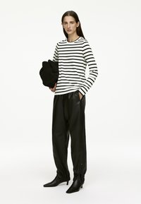 Schwarz-weiß gestreifte Langarmbluse, schwarze Lederhosen, spitze schwarze Absätze; das Model hält ein schwarzes Stoffstück. Minimalistischer Stil.