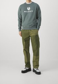 Hombre vistiendo un sudadera verde de Belstaff, pantalones cargo oliva de patchwork y zapatillas blancas con suela negra, de pie con las manos parcialmente en los bolsillos.