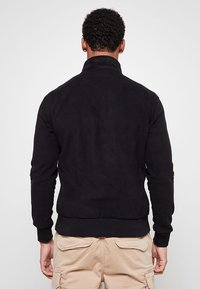 Giacca in pile nera con collo alto, maniche lunghe e fascia in vita a coste. Tessuto morbido con finitura liscia.