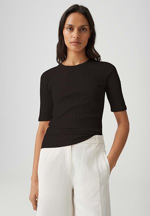 Vrouw staand met een neutrale uitdrukking, gekleed in een aansluitend zwart geribbeld top met korte mouwen en een witte broek met hoge taille.