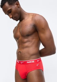 Μυώδης άνδρας που φοράει κόκκινα εσώρουχα Nike με λευκό λογότυπο, κοιτάζει προς τα κάτω, στέκεται μπροστά σε απλό ανοιχτό γκρι φόντο.