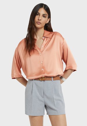 Frau mit langen dunklen Haaren, die ein locker sitzendes, pfirsichfarbenes Satin-Hemd mit Knöpfen und hoch taillierte, hellgraue, maßgeschneiderte Shorts mit braunem Gürtel trägt.