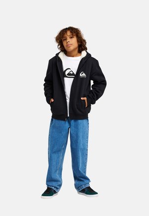 Boy with curly hair wearing black zip-up hoodie, white graphic t-shirt, blue jeans, and dark sneakers, standing with hands in pockets.