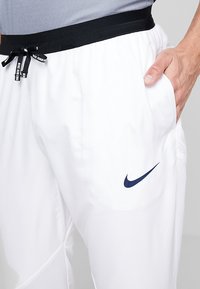 Osoba v bílých běžeckých šortkách Nike s černým pasem, s rukou odpočívající v pravé kapse, s logem Nike na stehně.