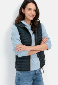 Gevoerd zwart vest over een lichtblauwe hoodie, met een ritssluiting aan de voorkant, contrasterende texturen en een decoratieve leren treklus.