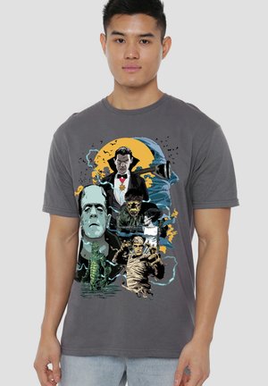 Man wearing a gray T-shirt with colorful graphic illustrations of classic movie monsters including Dracula, Frankenstein, the Mummy, and Creature from the Black Lagoon.