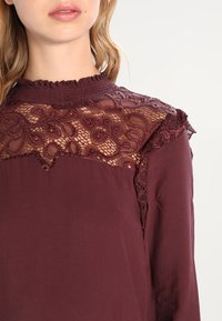 Blusa bordeaux con un'innesto in pizzo trasparente caratterizzato da disegni intricati e dettagli arricciati testurizzati sul colletto e sulle spalle.