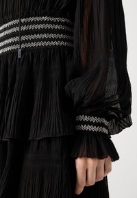 Black pleated dress with a ruffled design, featuring a textured waistband and embroidered silver accents on the cuffs. Soft, sheer fabric.