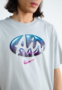 Világosszürke pamut póló, nagy, fényes, színes logóval, kék és lila árnyalatokkal, valamint egy kis rózsaszín Nike pipa logóval.